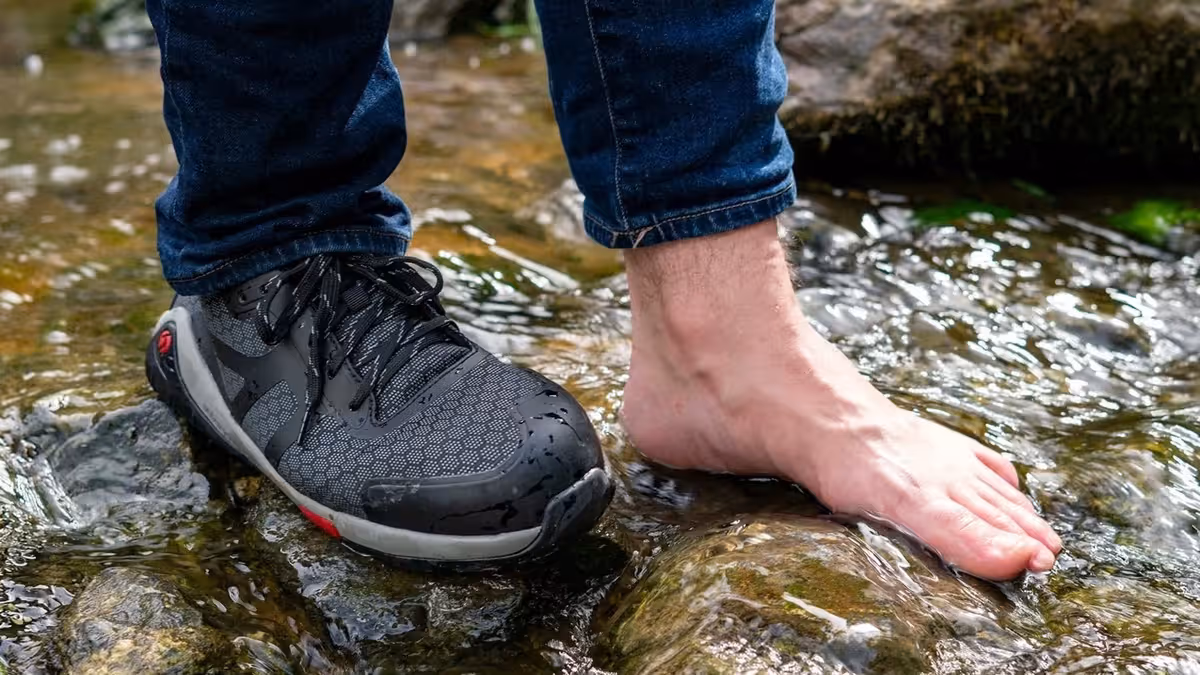 Trabajador con zapato de seguridad barefoot junto a un pie descalzo apoyado sobre rocas en un río
