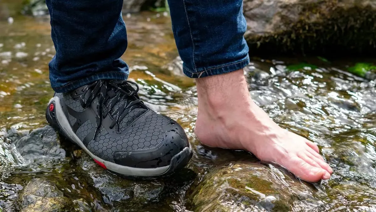 Trabajador con zapato de seguridad barefoot junto a un pie descalzo apoyado sobre rocas en un río