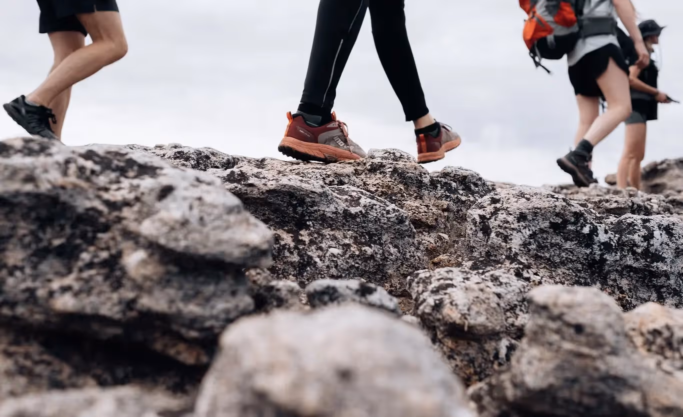 Tres peregrinos caminando por terreno rocoso de montaña con calzado deportivo de marcha; ilustra el cuidado de los pies durante una etapa larga del Camino de Santiago