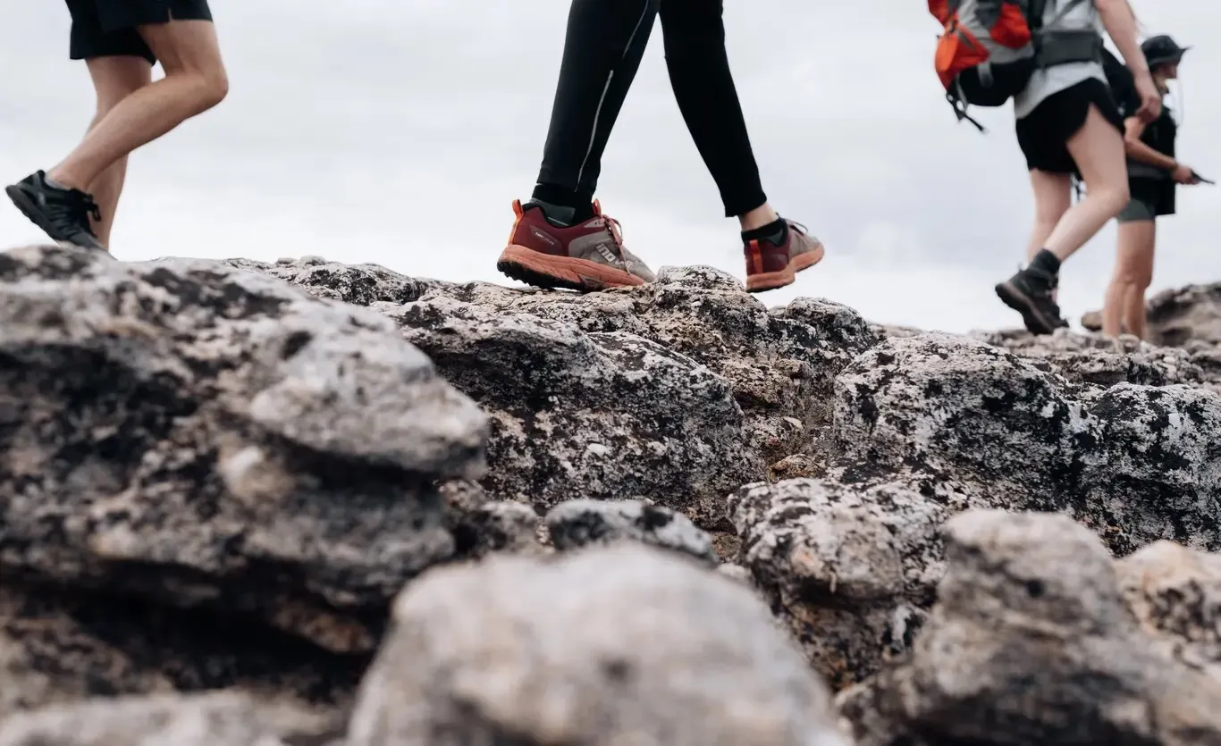 Tres peregrinos caminando por terreno rocoso de montaña con calzado deportivo de marcha; ilustra el cuidado de los pies durante una etapa larga del Camino de Santiago